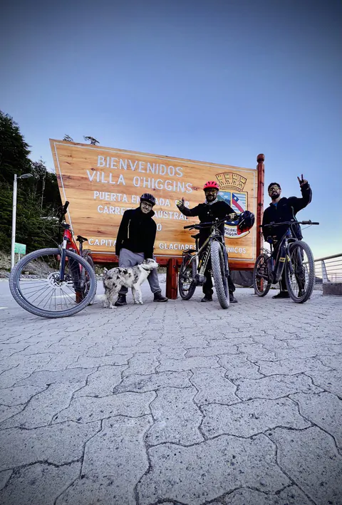 Tres riders con sus e-Bikes y un perro frente al cartel de bienvenida de Villa O'Higgins, puerta de entrada sur de la Carretera Austral