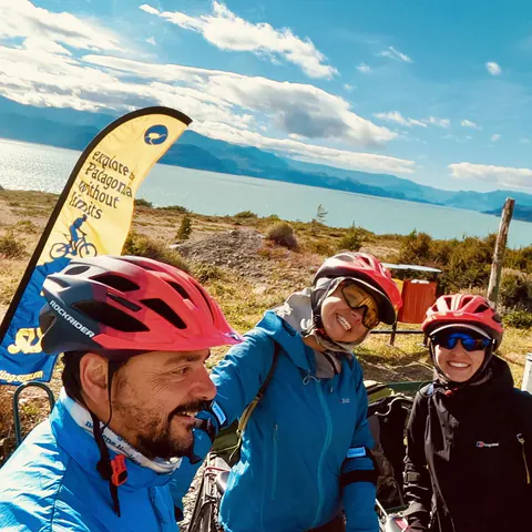 Dos riders y Javi, con cascos, frente al banderín de SURí y el lago O'Higgins, en Candelario Mancilla