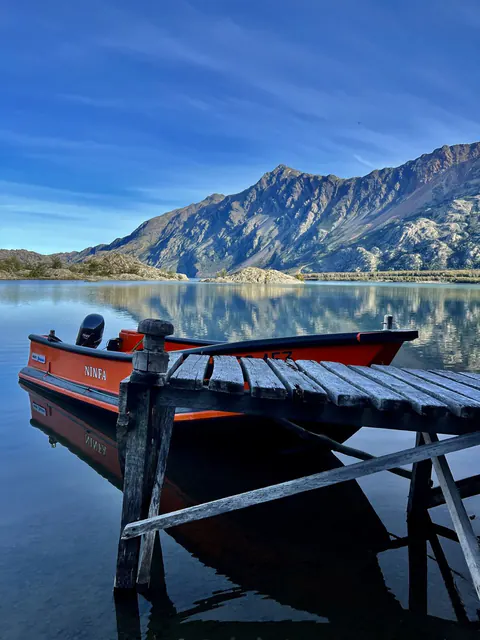 Un bote naranja amarrado a un muelle de madera, en un lago quieto bajo un cerro