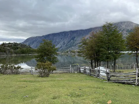 Un campo verde con cercos de palos y árboles, a orillas del lago, con cerros detrás