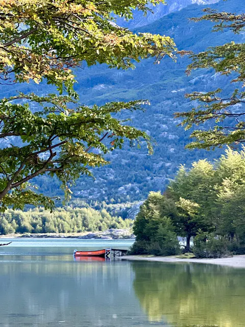 El lago Christie desde la orilla, entre árboles, con un bote rojo y cerros al fondo