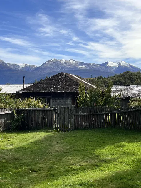 Una casa de campo con techo de tejuela y pasto verde, con cerros nevados detrás