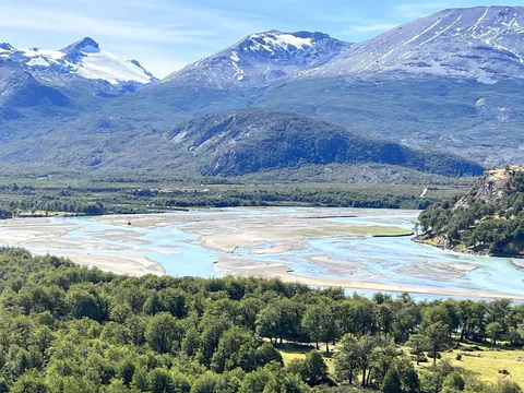 Un valle ancho con un río trenzado entre bosques, con cerros nevados al fondo