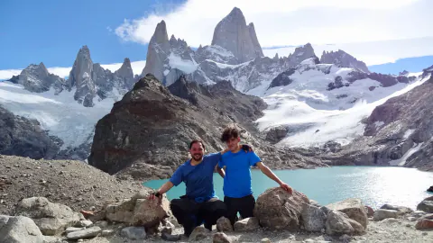 Dos personas sentadas sobre las rocas junto a una laguna, con el Fitz Roy y sus glaciares detrás