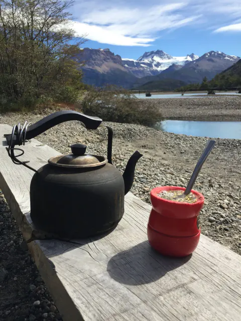 Una tetera negra y un mate rojo sobre una tabla de madera, frente a un lago y cerros nevados