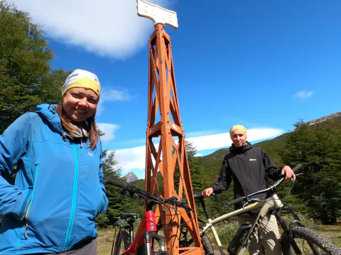 Dos riders junto a sus bicis al pie del hito fronterizo naranja del Paso Dos Lagunas, entre Chile y Argentina