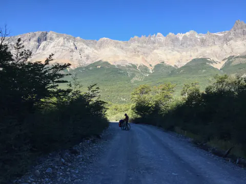 Un ciclista con casco y alforjas en un camino de ripio entre árboles, bajo una pared de cerros