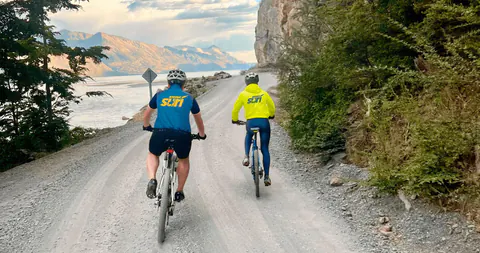 Dos riders con casco pedalean por el ripio de la Carretera Austral, entre la roca y el lago