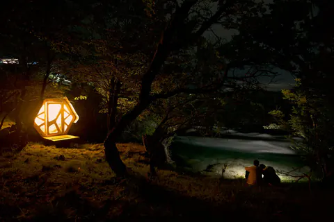 Un micro-refugio SURí iluminado de noche entre los árboles, junto al río Cochrane