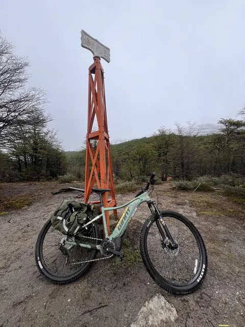 Una e-Bike con alforjas apoyada al pie del hito fronterizo de fierro que dice Chile