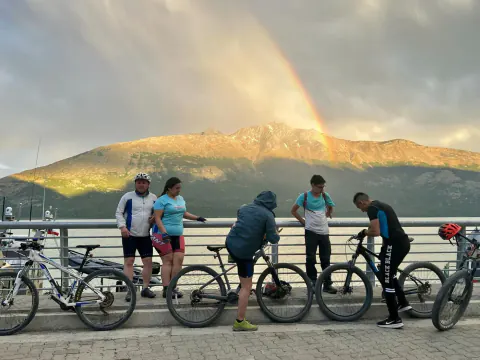 Cinco ciclistas con sus bicis en la costanera de Puerto Bahamondes, frente a un cerro iluminado y un arcoíris