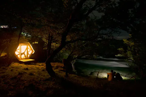 Un micro-refugio SURí iluminado de noche entre los árboles, junto al río Cochrane
